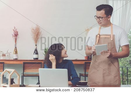 Young Asian man wear apron talking and receive order from customer at working table in coffee shop with smile face.Concept of cafe and coffee shop small business.