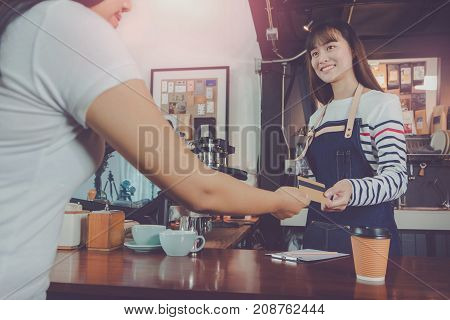 Young beautiful Asian woman barista wear blue apron and customer payment by credit card at bar counter in coffee shop with smile face.Concept of cafe and coffee shop small