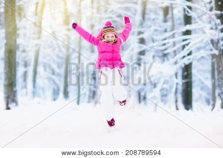Child playing with snow in winter. Little girl in colorful jacket and knitted hat catching snowflakes in winter park on Christmas. Kids play and jump in snowy forest. Children catch snow flakes