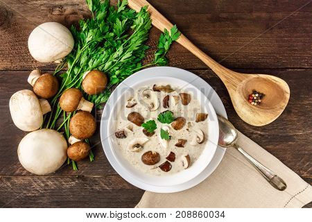 An overhead photo of a plate of a mushroom cream soup with champignons, cremini, fresh parsley, and peppercorns, on a dark rustic background texture with a place for text