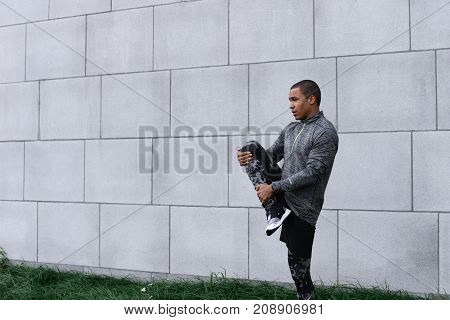 Picture of stylish slim young dark-skinned male jogger or runner wearing leggings running shoes and fleece jacket warming up legs outdoors before morning cardio workout routine. Sports and fitness