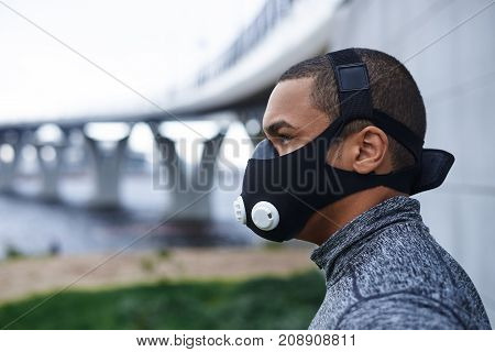 Profile picture of self-determined confident young African athlete wearing black training mask to increase load on breathing muscles during morning cardio workout routine outdoors. Sports and fitness