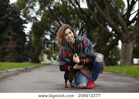 People pets leisure and activity. Cheerful young female in ripped jeans and warm plaid sitting on pavement on one knee embracing her faithful four legged friend. Owner and pet dog resting outdoors