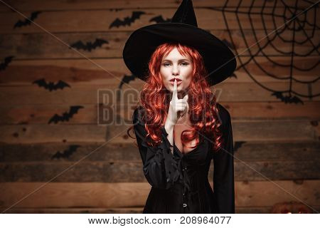 Halloween witch concept - Happy Halloween red hair Witch doing silence gesture with finger on her lips over old wooden studio background.
