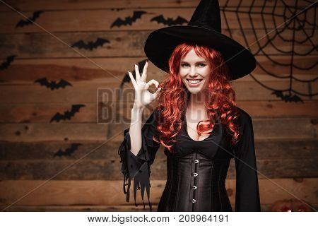 Halloween witch concept - Happy Halloween red hair Witch holding ok sign with fingers posing over old wooden studio background.