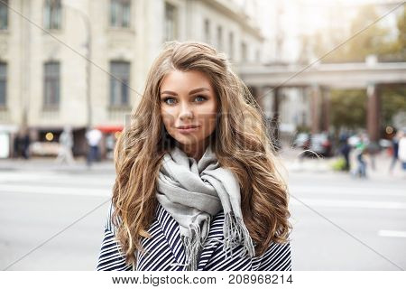Headshot of charming Caucasian young woman with light-brown wavy hair dressed in stripped coat and big grey scarf staring at camera with lazy smile standing on European city background