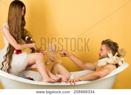 Love and romance. Woman in dress and guy with toy bear. Boyfriend and girlfriend relax on yellow background. Man in bathtub near girl with long hair. Couple in love of man and woman in bath.