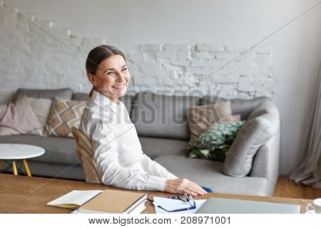 Picture of cheerful middle aged school psychologist having rest at her study sitting at desk with copybook notes and laptop computer. Beautiful senior female freelancer taking notes working at home