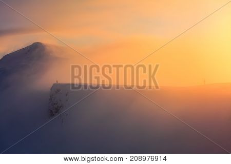 High in the mountains the sun is rising and enlightens everything with wonderful orange color. The mountains are in snow. Someone is staying at the edge of the rock. Unbelievable winter landscape.