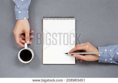 Woman's hands writing in notebook holding cup of coffee on gray workplace. Multitasking concept work and coffee break. Minimal creative playful. Top view.