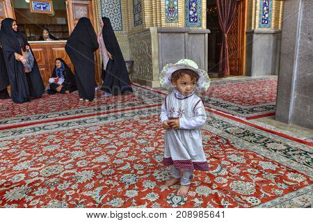 Shiraz Iran - 19 april 2017: Shah Cheragh Shrine An unknown little girl about 4 years old wearing a beach headdress stands at entrance to mosque near a group of women in black Muslim clothes.