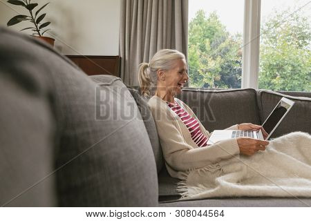 Side view of happy active senior Caucasian woman relaxing on sofa and using laptop in living room at comfortable home