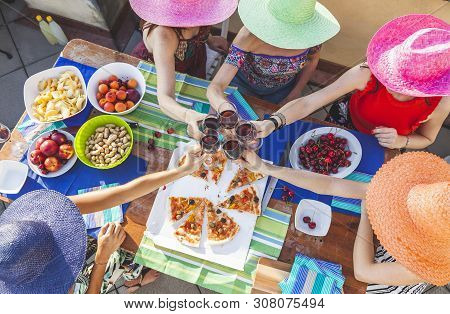 Group Of Female Friends Wearing Colorful Hats Toasting With Red Wine To Celebrate On A Terrace