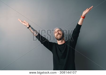 You Are Always Welcome. Handsome Young Man With Glasses, Beard And Tatooes, In Black Sweater Gesturi