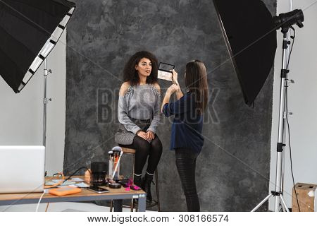 Portrait of female makeup artist applying cosmetics to pretty woman during photo shooting with professional camera and softbox in studio