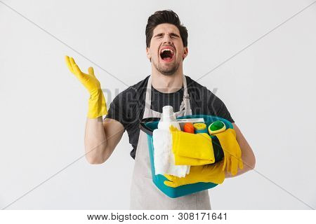 Handsome brunette houseman wearing apron standing isolated over white background, holding bucket full of detergents
