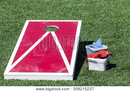 Red Corn Hole Game With White Stripes On A Green Turf Field With Blue And Red Bean Bags In Container