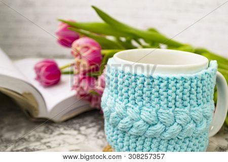 White Cup Of Tea In A Knitted Blue Case With An Open Book And With Pink Flowers Tulips And Sweets, W