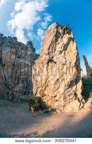 Athletic Man Climbs A Rock, 7c Category Route In The Forest With Rope, Against Sunset And The Other 