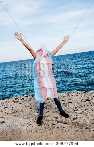 a young caucasian person covered with a transgender pride flag with his or her arms open in the air, with the ocean in the background