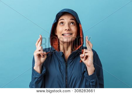 Cheerful young girl wearing raincoat standing isolated over blue background, posing, fingers crossed