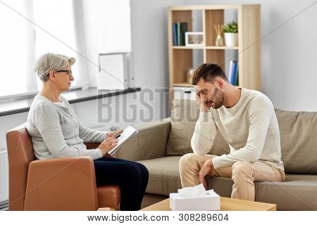psychology, mental therapy and problem concept - senior woman psychologist taking notes to notebook and sad young man patient at psychotherapy session