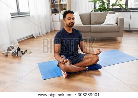 fitness, meditation and healthy lifestyle concept - indian man meditating in lotus pose on exercise mat at home