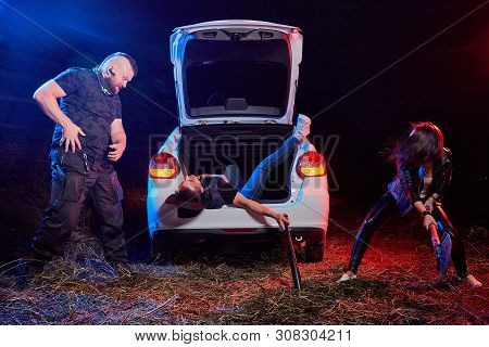 Couple Of Thugs Near Car And Dead Young Guy In The Trunk At Night. Photoshoot About Life Of Gungster