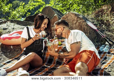 Young Couple Having Picnic At Riverside In Sunny Day. Woman And Man Spending Time On The Nature Toge
