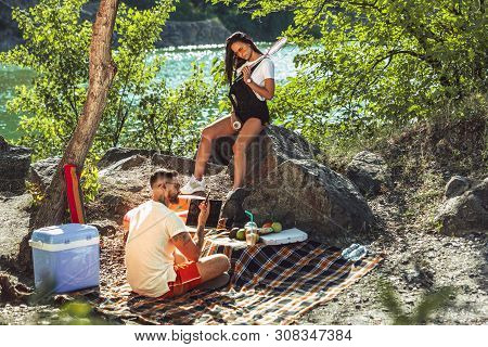 Young Couple Having Picnic At Riverside In Sunny Day. Woman And Man Spending Time On The Nature Toge