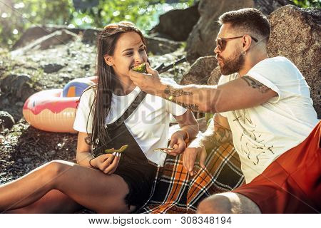 Young Couple Having Picnic At Riverside In Sunny Day. Woman And Man Spending Time On The Nature Toge