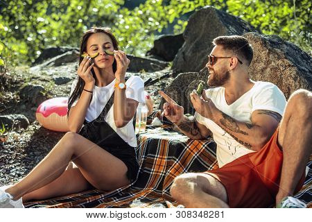 Young Couple Having Picnic At Riverside In Sunny Day. Woman And Man Spending Time On The Nature Toge