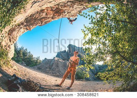 Athletic Man Climbs A Rock, 7c Category Route In The Forest With Rope, Against Sunset And The Other 