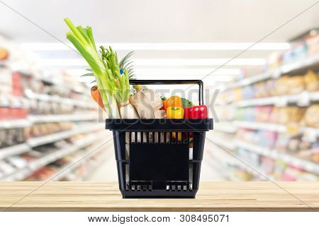 Food And Groceries In Black Shopping Basket On Wood Table With Blurred Suppermarket Aisle In Backgro