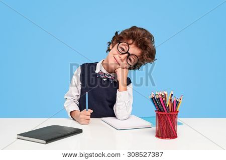 Adorable Smart Kid In Round Glasses Smiling And Supporting Head While Sitting At Desk During Studies