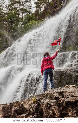 Tourist Woman With Norwegian Flag At Svandalsfossen In Norway, Powerful Waterfall In Norwegian Mount