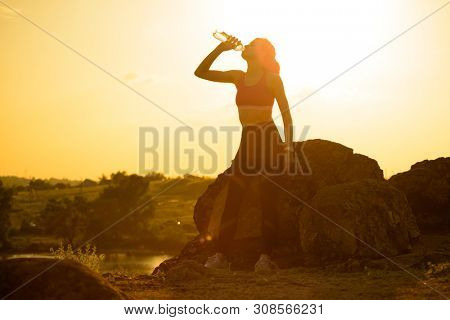 Young Woman Resting and Drinking Water after Trail Running Outdoor. Workout Exercise at Hot Beautiful Summer Sunset. Sport and Healthy Active Lifesyle Concept.