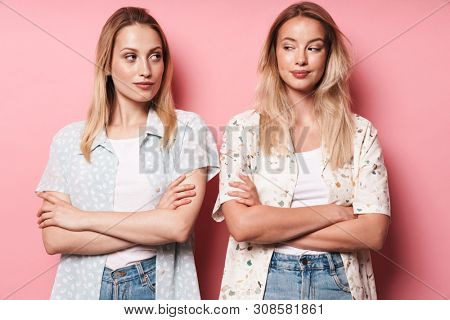 Two attractive sneaky blonde girls wearing summer outfit standing isolated over pink background, arms folded, looking at each other