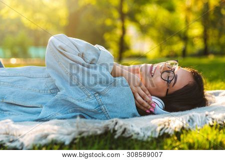 Image of a pleased smiling cute young student girl wearing eyeglasses outdoors in nature park listening music with heaphones lies on grass.