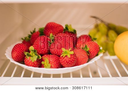 Plate With Ripe Strawberries Is On The Shelf Of The Refrigerator, Next To A Lemon And Grapes. The Co