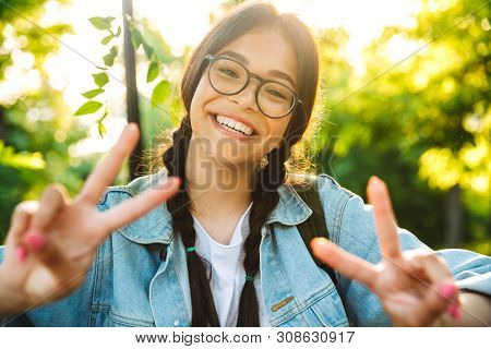 Image of a smiling cute young student girl wearing eyeglasses outdoors in nature park make peace gesure.