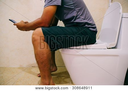 Using A Mobile Phone On Toilet. The Asian Man Sitting In The Toilet And Working Or Play Game In Phon