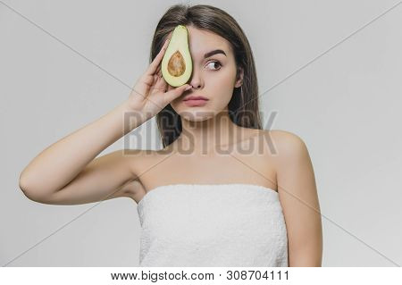 Young Astonished Girl With Half Avocado Is Looking Leftward. She Wears White Towel.