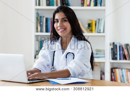 Laughing Hispanic Female General Practitioner Working On Computer At Office Of Hospital