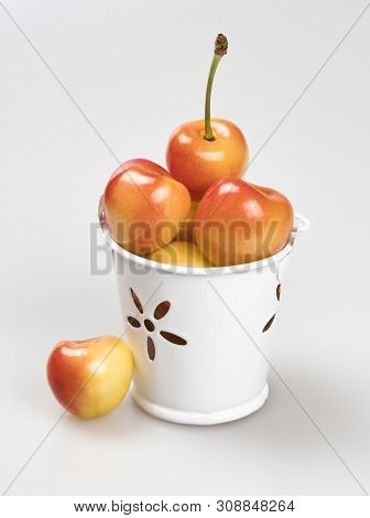 Berries Of Yellow-pink Cherries In A White Metal Bucket And One Near It, Close-up On A White Backgro