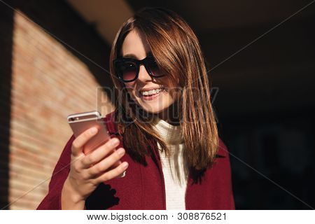 Young Smiling Lady In Sunglasses Standing On Street And Happily Using Her Mobile Phone
