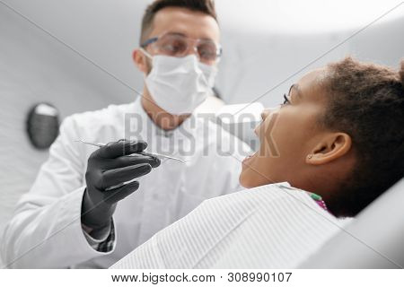 Side View Of African Girl Lying On Dentist Chair With Open Mouth While Doctor In Mask And Glasses Ke