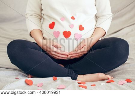 Closeup Of Pregnant Woman In White Clothes Sitting On Bed Touching Her Belly. Pregnant Belly Covered