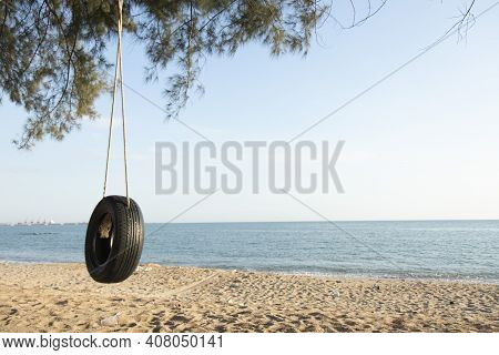 Tire Swings Hang From Trees On The Sandy Beach. With A Background Of Blue Sea And Sky With Coluds