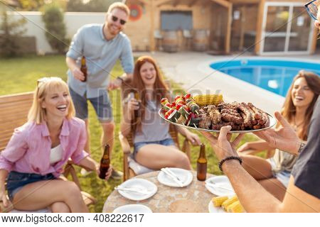 Group Of Cheerful Young Friends Gathered Around The Table, Drinking Beer And Having Fun At Backyard 